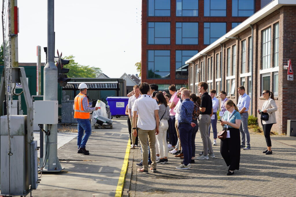 A group of people standing in a courtyard where railway technologies are displayed. A person in a reflective vest and helmet is presenting in front of them.