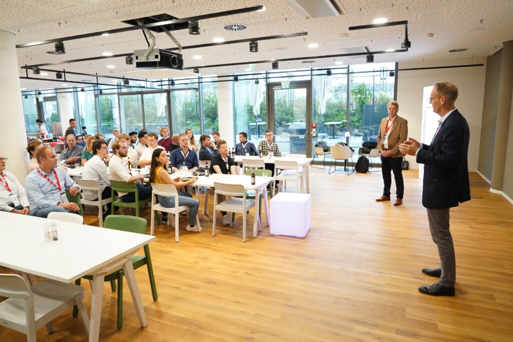 Two people standing in front of a large audience presenting. The presenters are: Karel van Gils and Volker Hentschel.