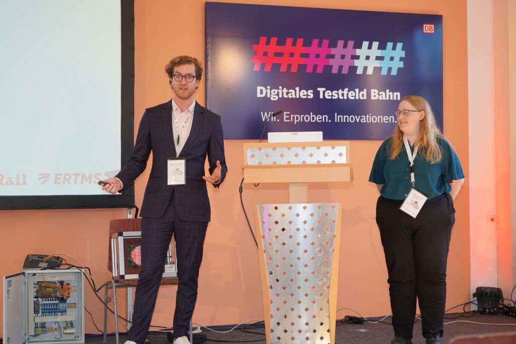 Two people presenting on a stage. In the background there is a sign saying "Digitales Testfeld Bahn. Wir. Erproben. Innovationen."